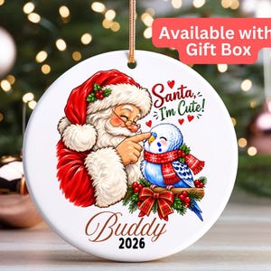 May include: A round white ceramic ornament featuring a colourful illustration of Santa Claus touching the beak of a blue budgie wearing a red scarf. The text reads Santa, I'm Cute! and Buddy 2026. The ornament hangs from a gold string.
