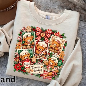 May include: A sand-coloured crewneck sweatshirt featuring a festive graphic of four framed illustrations of teddy bears wearing chef hats and baking cookies, surrounded by holly, candy canes, gingerbread men, and a red and white chequered bow.