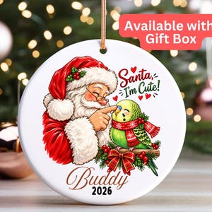 May include: A round white ceramic ornament featuring a graphic of Santa Claus touching the beak of a green budgie wearing a red scarf. The text reads Santa, I'm Cute! and Buddy 2026. The ornament has a gold hanging string at the top.
