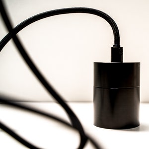 May include: A black cylindrical pendant light fixture with a black braided cord. The fixture has a matte finish and a simple, modern design. The cord curves gracefully against a white background.