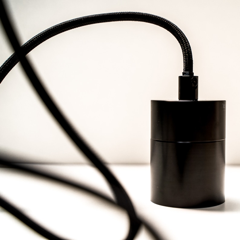 May include: A black cylindrical pendant light fixture with a black braided cord. The fixture has a matte finish and a simple, modern design. The cord curves gracefully against a white background.