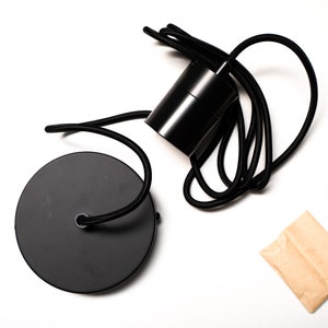 May include: Black pendant light fixture with a round ceiling plate and cylindrical housing. The fixture has a black fabric-covered cord. The components are on a white background.