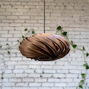 K&ouml;nnte beinhalten: Eine Holz-Pendelleuchte mit einem einzigartigen Spiraldesign. Die Leuchte besteht aus d&uuml;nnen Holzstreifen, die &uuml;bereinander gelegt und gebogen sind, um einen wirbelnden Effekt zu erzeugen. Die Leuchte h&auml;ngt an einem schwarzen Kabel vor einer wei&szlig;en Backsteinwand.