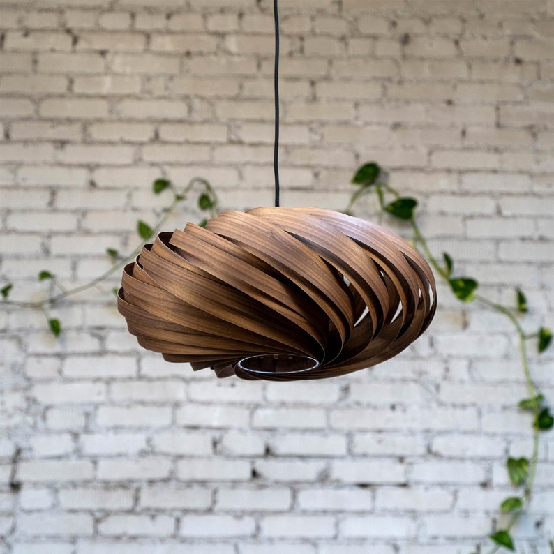 K&ouml;nnte beinhalten: Eine Holz-Pendelleuchte mit einem einzigartigen Spiraldesign. Die Leuchte besteht aus d&uuml;nnen Holzstreifen, die &uuml;bereinander gelegt und gebogen sind, um einen wirbelnden Effekt zu erzeugen. Die Leuchte h&auml;ngt an einem schwarzen Kabel vor einer wei&szlig;en Backsteinwand.