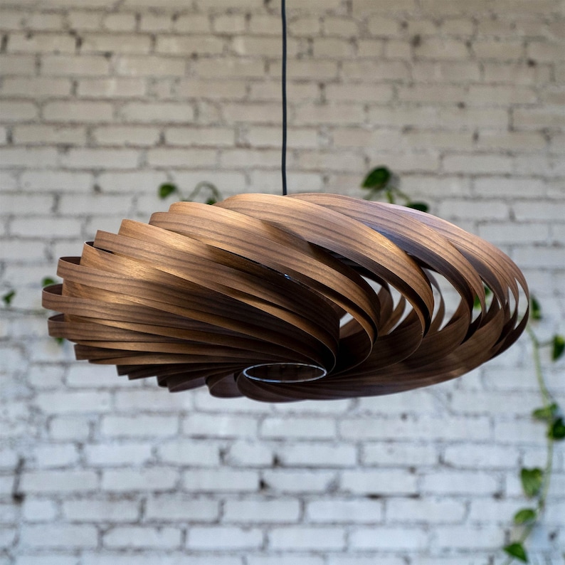 May include: A modern, wooden pendant light fixture with a spiral design. The light is suspended from a black cord and features a warm, brown wood finish. The fixture is set against a white brick wall.