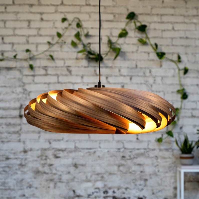 May include: A modern, wooden pendant light fixture. The light has a spiral design made of layered wood, with warm light emanating from within. The fixture hangs from a black cord against a white brick wall.