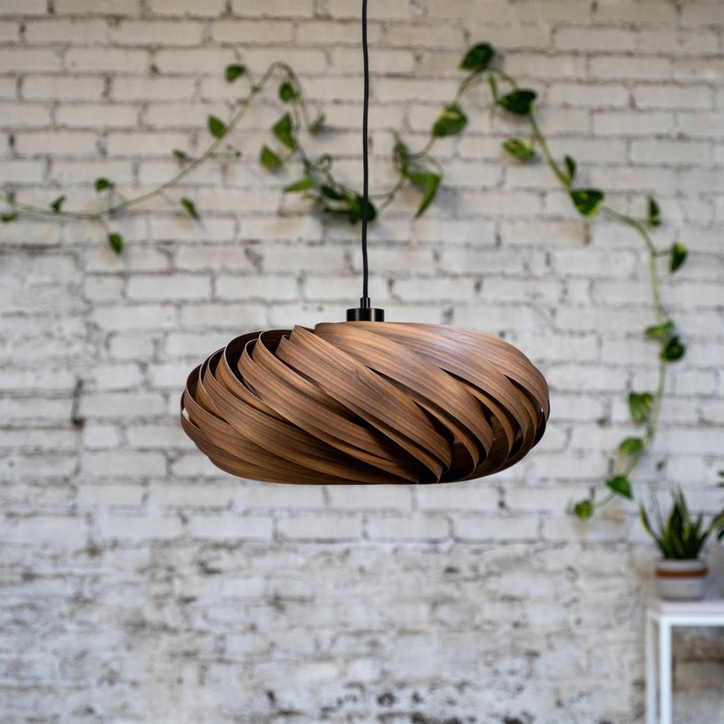K&ouml;nnte beinhalten: Eine Holz-Pendelleuchte mit einem einzigartigen, wirbelnden Design. Die Leuchte besteht aus d&uuml;nnen Holzstreifen, die &uuml;bereinander gelegt und gebogen sind, um einen modernen und organischen Look zu erzeugen. Die Leuchte h&auml;ngt an einem schwarzen Kabel vor einer wei&szlig;en Backsteinwand.