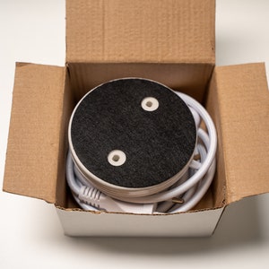 May include: A white round base with a black top and two small holes. The base is in a cardboard box with a white lid. A white cord is wrapped around the base.