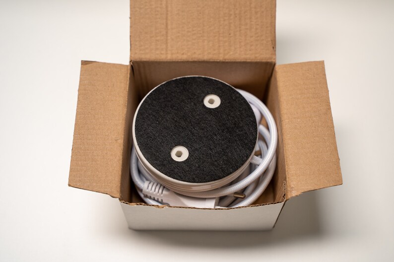 May include: A white round base with a black top and two small holes. The base is in a cardboard box with a white lid. A white cord is wrapped around the base.