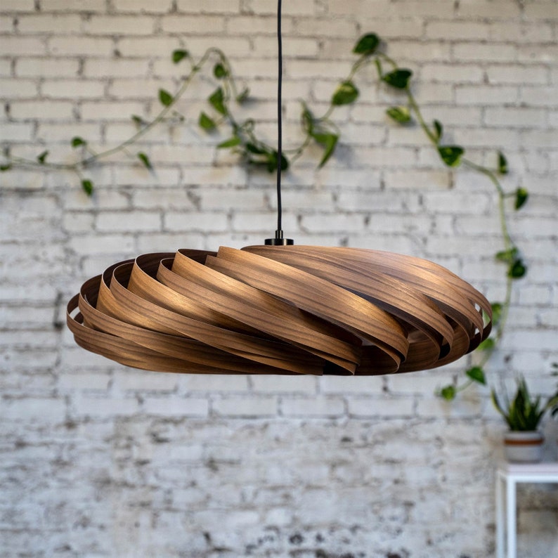 May include: A wooden pendant light fixture with a spiral design. The light is suspended from a black cord and features a warm brown color. The fixture is set against a white brick wall with green vines.