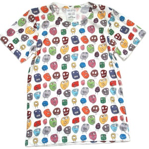Pode incluir: Camiseta branca de manga curta com um padrão colorido de rostos de desenhos animados. O texto "Camp Meadows" é impresso na camiseta.