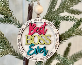 May include: A round wooden ornament with the words "Best Boss Ever" in colorful letters. The ornament is decorated with a circle of words describing positive qualities of a boss, such as "caring", "honest", "supportive", "decisive", "trustworthy", "compassionate", "inspirational", "motivational".