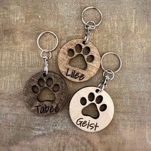 May include: Three wooden keychains with a paw print design and personalized names. The keychains are brown and have silver key rings. The names on the keychains are "Lilee", "Tabee", and "Geist".