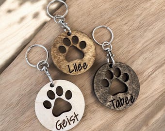 May include: Three circular wooden keychains with silver metal rings and lobster clasps. Each keychain features a cutout paw print design and a laser-engraved name. The names Lilee, Tabee, and Geist are printed in a dark script font on the wood.