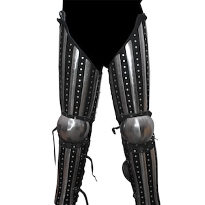 May include: Pair of medieval-style leg armor. The armor is primarily black with vertical silver stripes and rivets. The knee caps are rounded and silver. Leather straps and ties are visible.