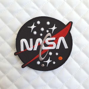 Peut inclure: Écusson brodé avec le logo de la NASA. Le patch circulaire a un fond noir avec le logo de la NASA en blanc. Un trait rouge et des étoiles blanches sont également présents. Le patch est destiné à être appliqué sur des vêtements ou des accessoires.
