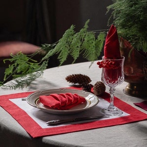 May include: A red placemat with a white border and a white linen tablecloth. A white plate with a red napkin folded on top sits on the placemat. A glass of water and a small pine cone are on the table.