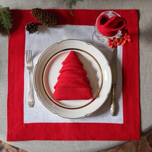 May include: A festive Christmas table setting with a red and white placemat. A white plate with a gold rim holds a red napkin folded into a Christmas tree shape. Silverware and a glass with a folded napkin and berries complete the scene.