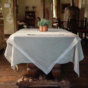 Puede incluir: Un mantel de mesa de lino gris claro con ribete de encaje blanco, drapeado sobre una mesa de madera con patas talladas. Una pequeña planta en maceta se encuentra sobre la mesa.