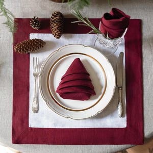 May include: A white linen placemat with a burgundy border, a white plate with a gold rim, a burgundy napkin folded into a Christmas tree shape, a silver fork and knife, and pine cones on a beige tablecloth.
