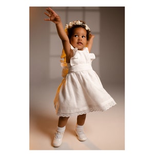 Può includere: Una bambina che indossa un vestito bianco con un colletto a balze e una corona di fiori. È in piedi su una superficie marrone chiaro e ha le braccia alzate sopra la testa.