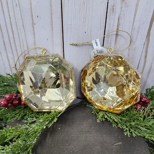 May include: Two gold-colored glass ornaments shaped like diamonds. The ornaments have a sparkly finish and are hanging from gold string.
