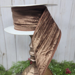 May include: A spool of brown velvet ribbon with a stitched edge. The ribbon is a rich, warm brown color and has a soft, textured appearance. The ribbon is displayed on a white cardboard spool.