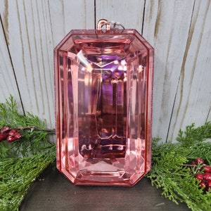 May include: A large, pink, faceted ornament in a rectangular shape with a glittery pink border. The ornament has a copper-colored loop at the top for hanging. Greenery and red berries are in the foreground.