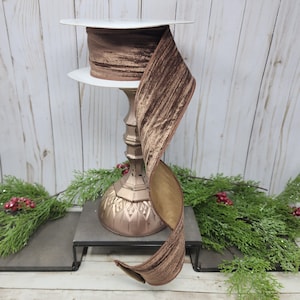May include: A spool of brown velvet ribbon with a textured surface. The ribbon is displayed on a decorative gold-colored stand. The ribbon is a rich brown color with a lighter brown reverse side. The ribbon is suitable for crafts and gift wrapping.