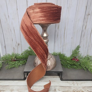 May include: A roll of wide, rust-colored velvet ribbon, partially unrolled, sits atop a decorative silver candlestick. The ribbon has a soft, textured appearance and is displayed against a light wood-paneled background. The ribbon is approximately 2 inches wide.