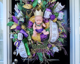 May include: A Mardi Gras wreath with a baby doll wearing a purple outfit and a gold crown. The wreath is decorated with purple, green, and gold beads, ribbons, and feathers. It also has a sign that says "Mardi Gras".