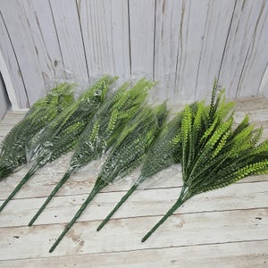 May include: A bunch of six artificial green fern sprigs in plastic packaging. The sprigs are bundled together and have long stems.
