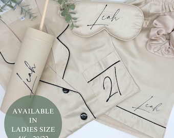 Op de afbeelding: Een beige satijnen pyjamaset met zwarte bies, een bijpassend slaapmasker, scrunchie en beker. De set bevat een overhemd met knopen en een korte broek. De naam "Leah" staat op de beker, het slaapmasker en de korte broek. Het nummer "21" staat op de zak.