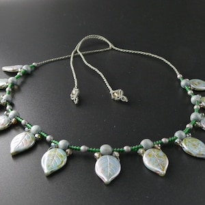 Op de afbeelding: Een zilveren en groene kralenketting met een reeks zilveren bladvormige hangers.