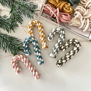 Puede incluir: Un conjunto de cinco adornos de bastón de caramelo DIY hechos con cuerda de algodón en varios colores, incluyendo azul verdoso, rosa, amarillo, verde y blanco. Los adornos están empaquetados en una caja con carretes de cuerda de algodón en diferentes colores.