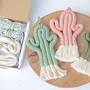 May include: Three macrame cactus wall hangings in green, pink, and olive green colours. Each cactus has a white tassel at the bottom. The cactus hangings are displayed on a wooden surface.