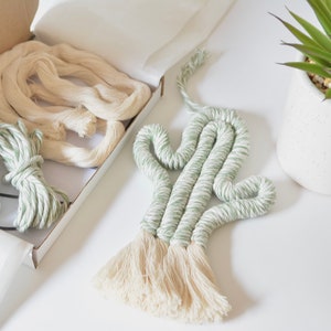 Op de afbeelding: Een groene en witte macramé cactus wandhanger met een crèmekleurige kwast. De cactus zit in een doos met twee spoelen groene en witte draad en een spoel crèmekleurige draad.