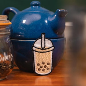 May include: A blue ceramic teapot with a matching lid and a small, embroidered patch of a bubble tea drink. The patch features a beige drink with brown boba pearls and a white lid. A glass jar is visible to the left.