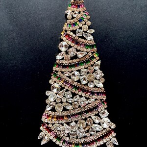 May include: A decorative Christmas tree ornament crafted from numerous clear, white, and multi-coloured rhinestones. The tree features rows of baguette-cut stones in red, green, and purple, interspersed with various shapes of clear, faceted crystals.