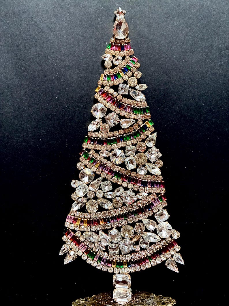 May include: A decorative Christmas tree ornament crafted from numerous clear, white, and multi-coloured rhinestones. The tree features rows of baguette-cut stones in red, green, and purple, interspersed with various shapes of clear, faceted crystals.