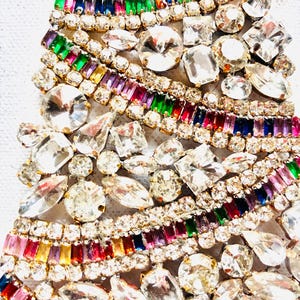 May include: A decorative object shaped like a Christmas tree, densely covered in clear, faceted rhinestones and rows of rectangular, multi-coloured crystals in shades of red, green, blue, purple, and yellow, all set in a gold-toned metal frame.