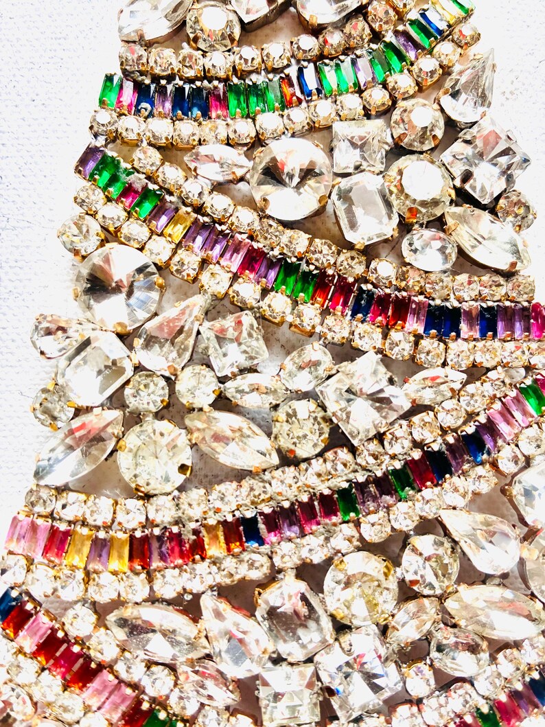 May include: A decorative object shaped like a Christmas tree, densely covered in clear, faceted rhinestones and rows of rectangular, multi-coloured crystals in shades of red, green, blue, purple, and yellow, all set in a gold-toned metal frame.