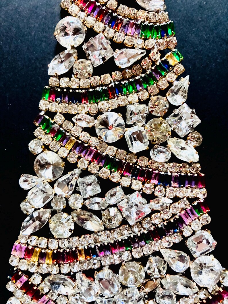May include: A decorative brooch shaped like a Christmas tree, constructed from numerous clear, round, pear, and emerald-cut rhinestones alongside rows of multi-coloured baguette-cut crystals in shades of red, green, blue, purple, and yellow.