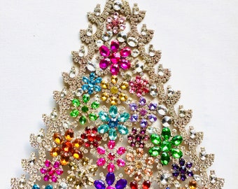 May include: A colorful Christmas tree made of rhinestones. The tree is decorated with various colors of rhinestones, including red, blue, green, yellow, and purple. The tree is standing on a gold base.