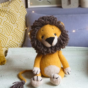 May include: A knitted lion toy with a yellow body, white belly, and brown mane. The toy is sitting on a light green blanket with fringe.