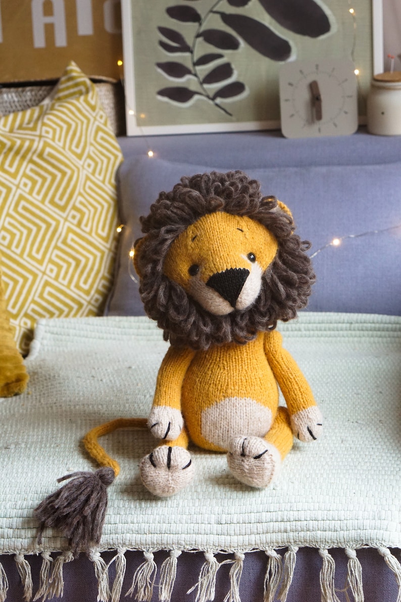 May include: A knitted lion toy with a yellow body, white belly, and brown mane. The toy is sitting on a light green blanket with fringe.