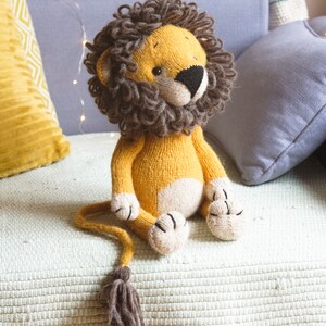 May include: A knitted lion toy with a brown mane, yellow body, and white paws. The toy is sitting on a light green blanket with fringe.