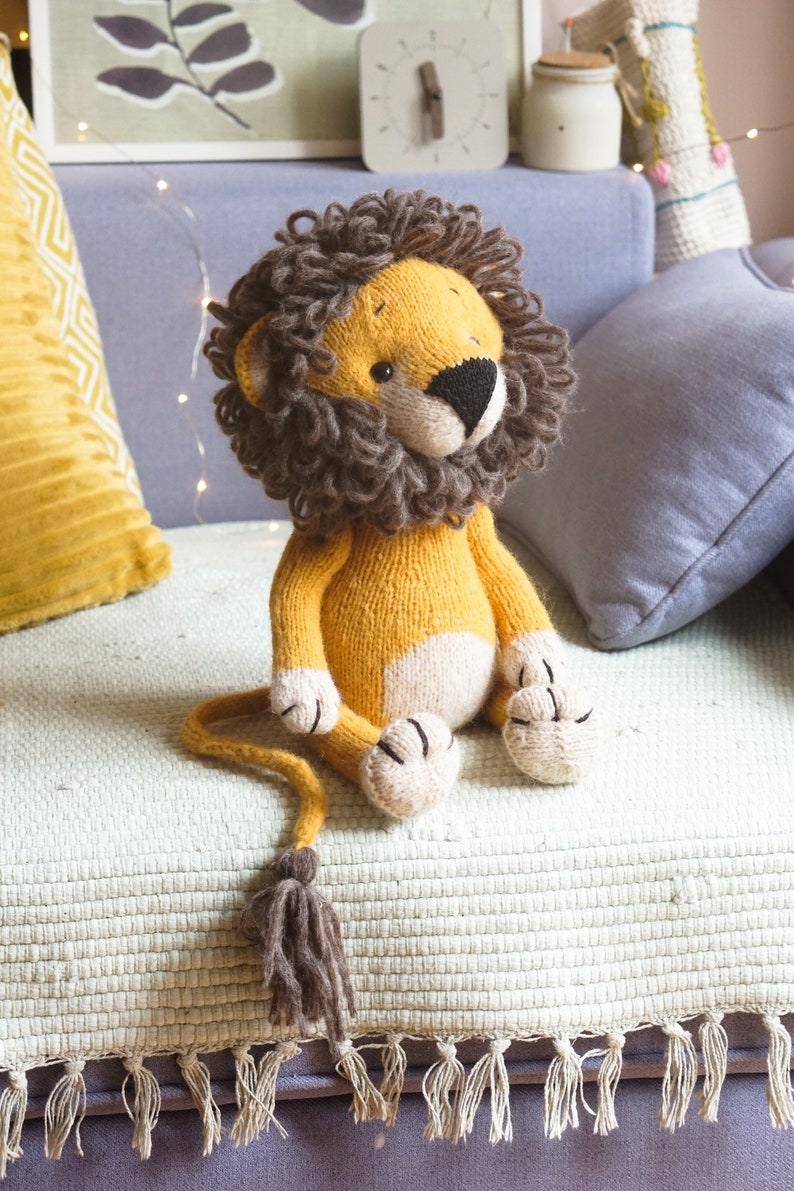 May include: A knitted lion toy with a brown mane, yellow body, and white paws. The toy is sitting on a light green blanket with fringe.