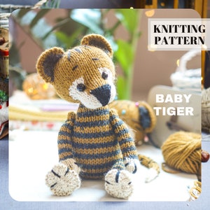 May include: A knitted baby tiger toy with a tan and navy blue striped body, white face, and black nose. The ears and paws are tan with dark gray accents. The text "KNITTING PATTERN" and "BABY TIGER" are visible.