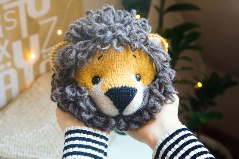 May include: A knitted lion head with a brown mane and a yellow face. The lion has black eyes and a black nose.